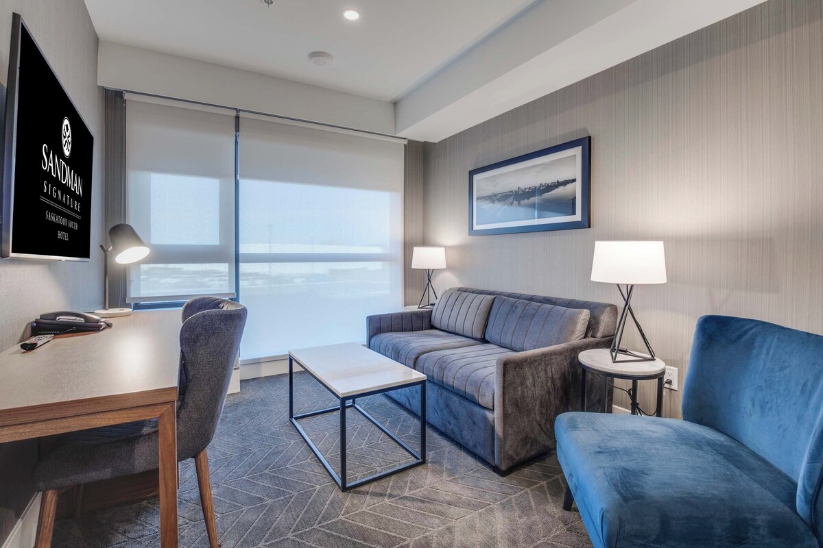 A modern hotel room with a sofa, armchair, desk, and lamps.