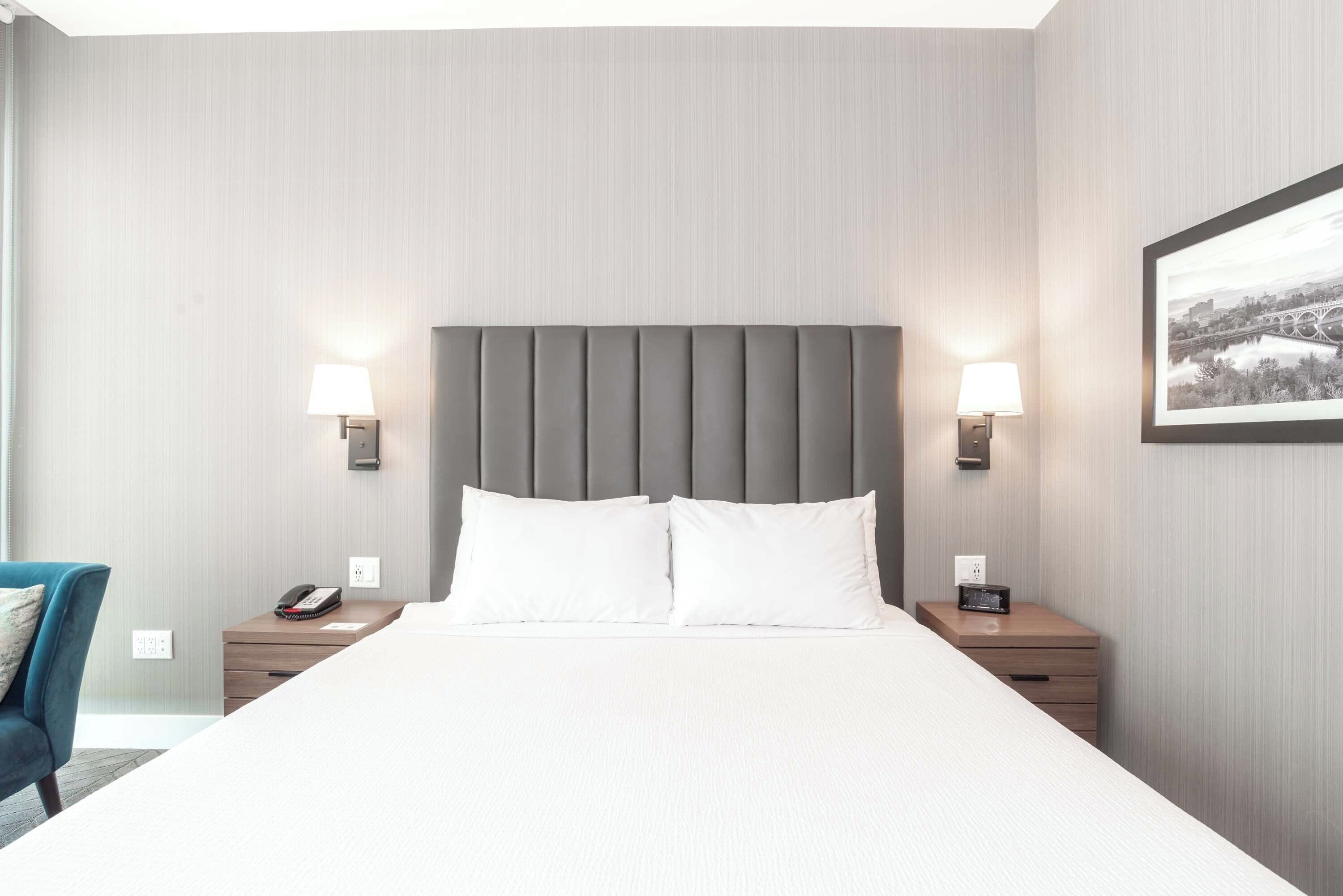 A hotel room with a large bed, two bedside tables with lamps, a chair, and a framed picture on the wall.