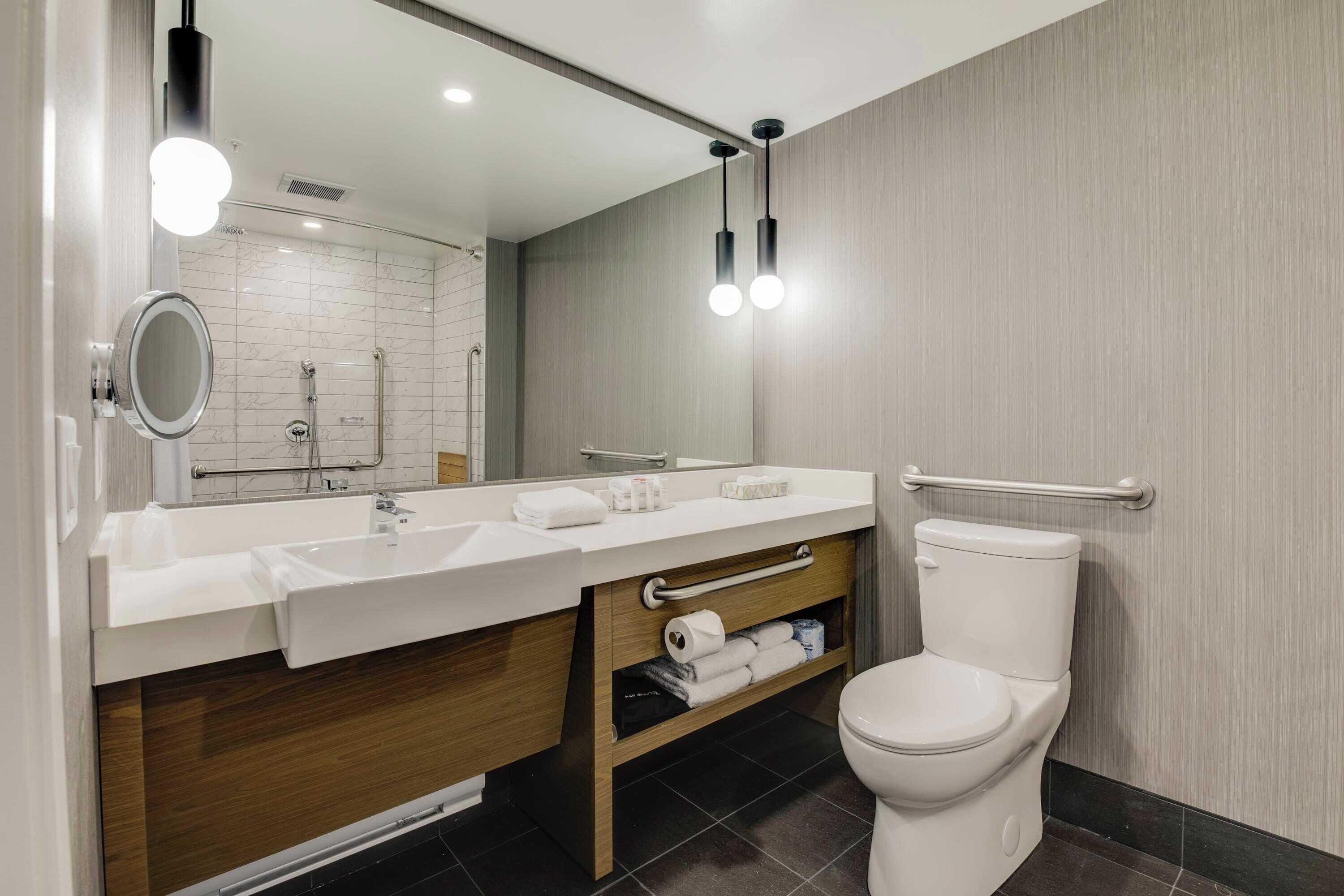 Une salle de bain moderne avec un lavabo blanc, un miroir et des toilettes. Il y a deux luminaires au plafond et un porte-serviettes.