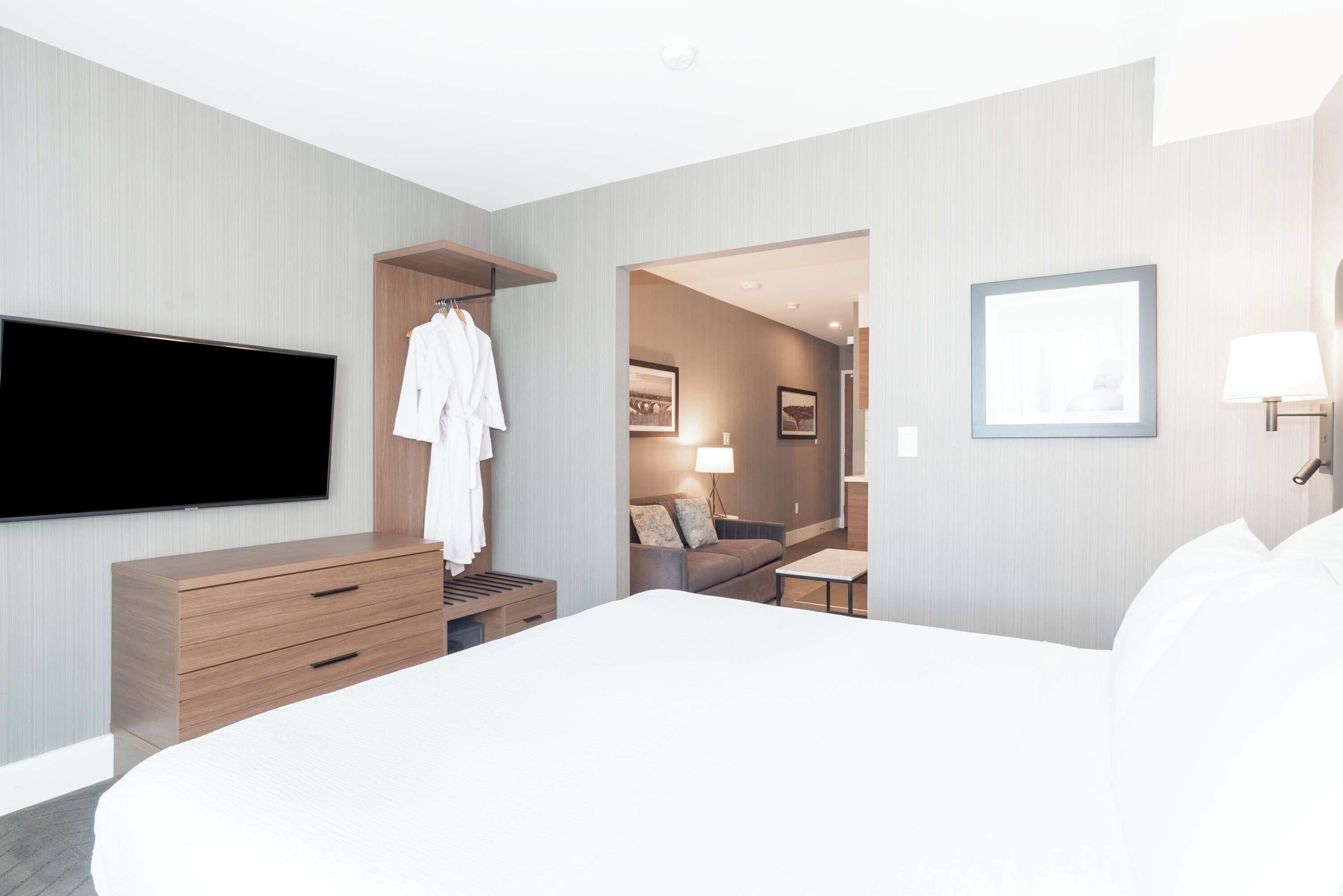A modern hotel room with a large bed, a TV mounted on the wall, a wooden dresser, and a walk-in closet with a robe hanging.