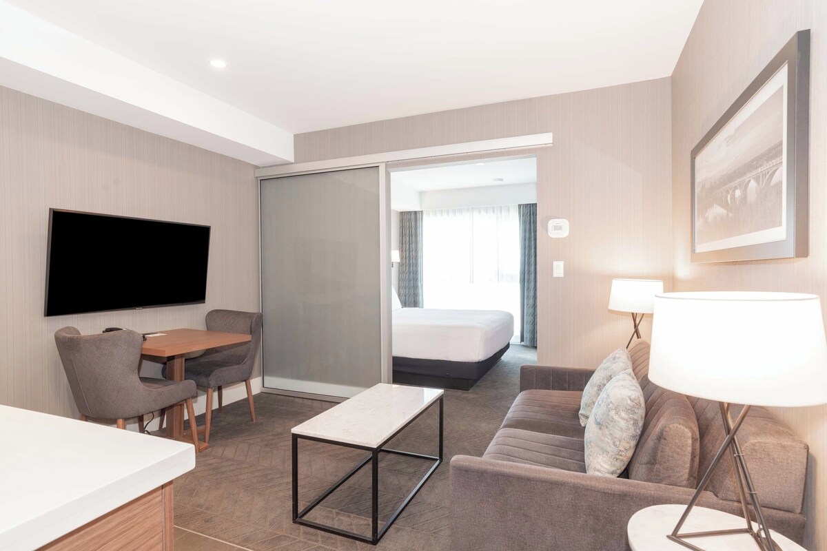 A modern hotel room with a flat-screen TV, a sofa, a coffee table, and a bed with a sliding door leading to a balcony.