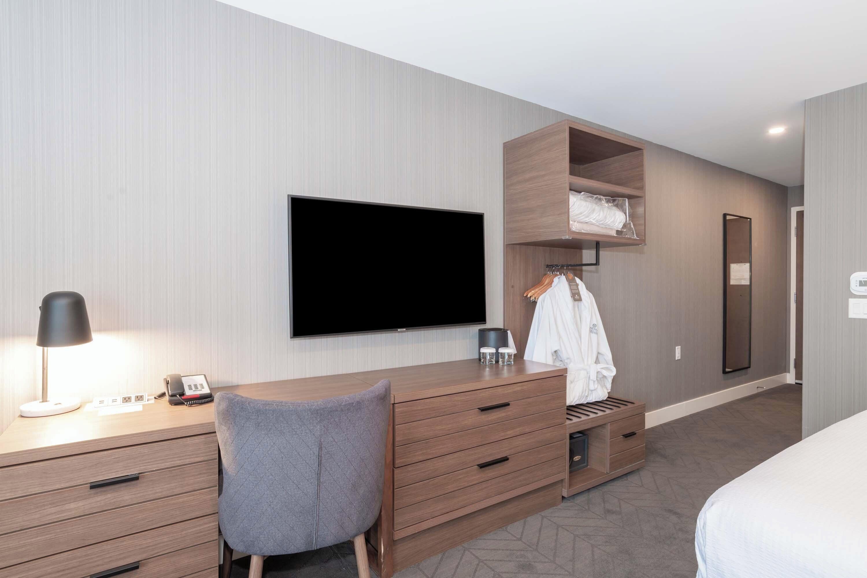 A modern hotel room with a wooden desk, a flat-screen TV, a grey chair, a wardrobe, and a bed.