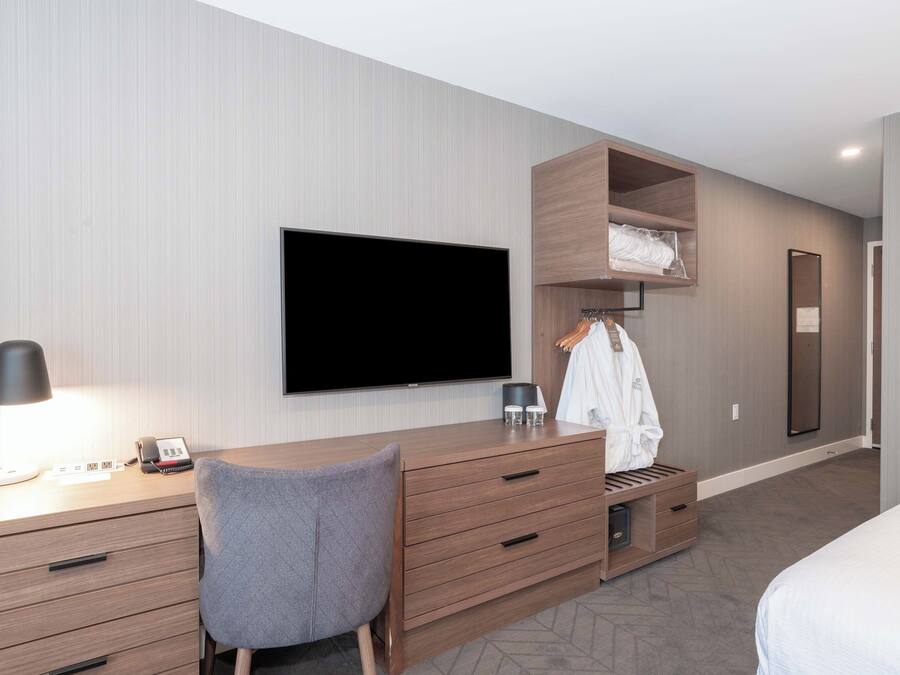 A modern hotel room with a wooden desk, a flat-screen TV, a grey chair, a wardrobe, and a bed.