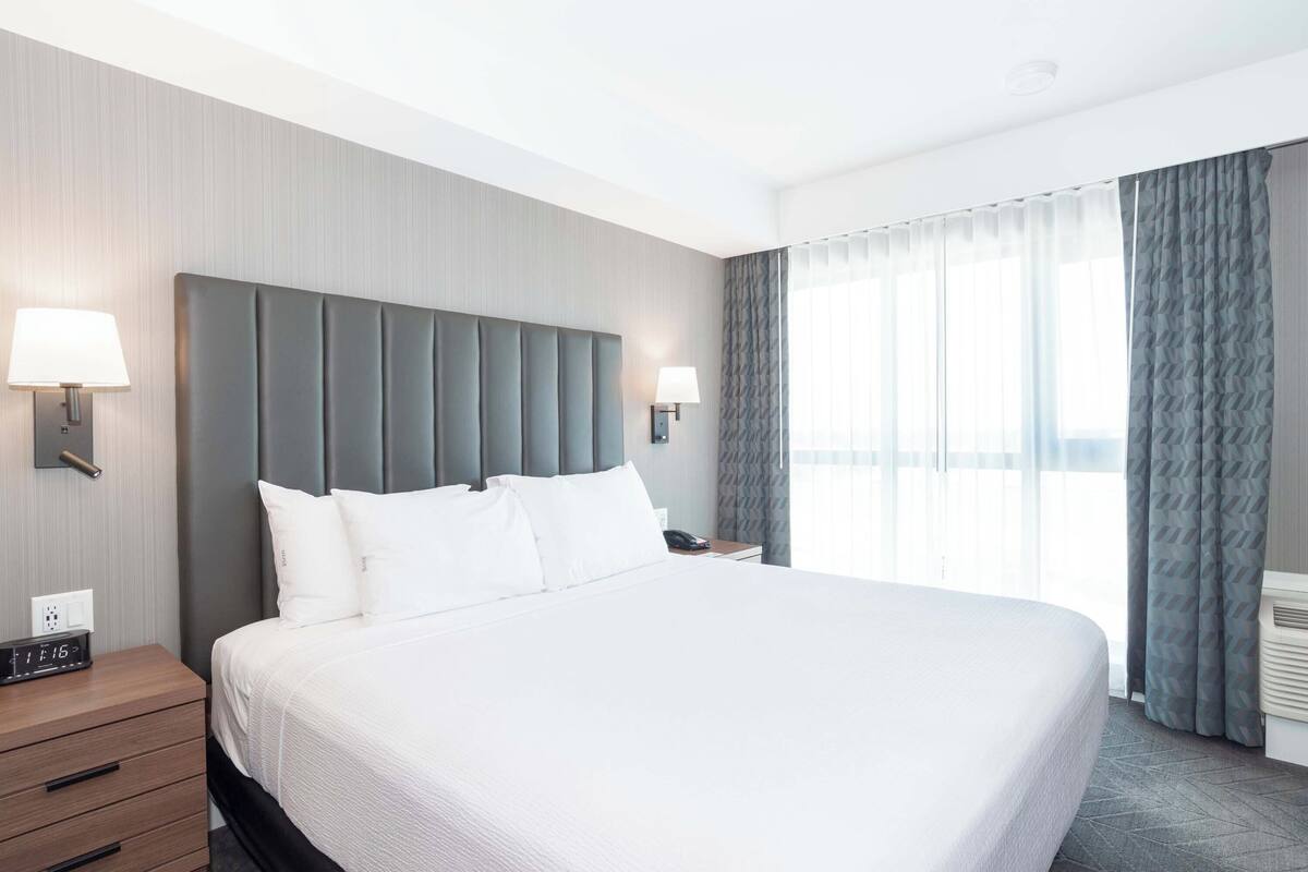 Habitación de hotel con una cama grande, una mesita de noche con lámpara, un reloj de pared y una ventana con cortinas.