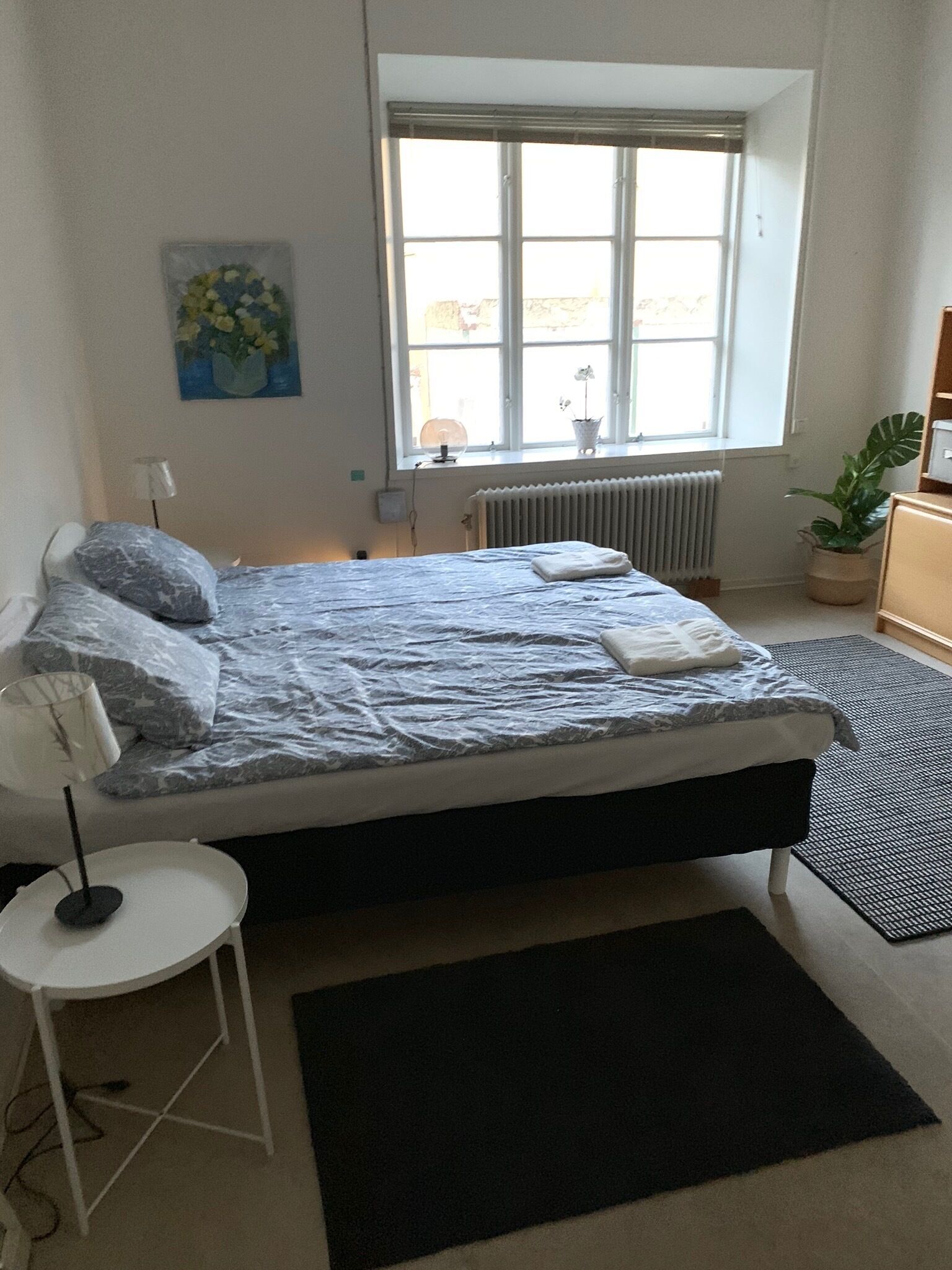 Un dormitorio con cama, una mesita, un radiador, una ventana y una planta en maceta.