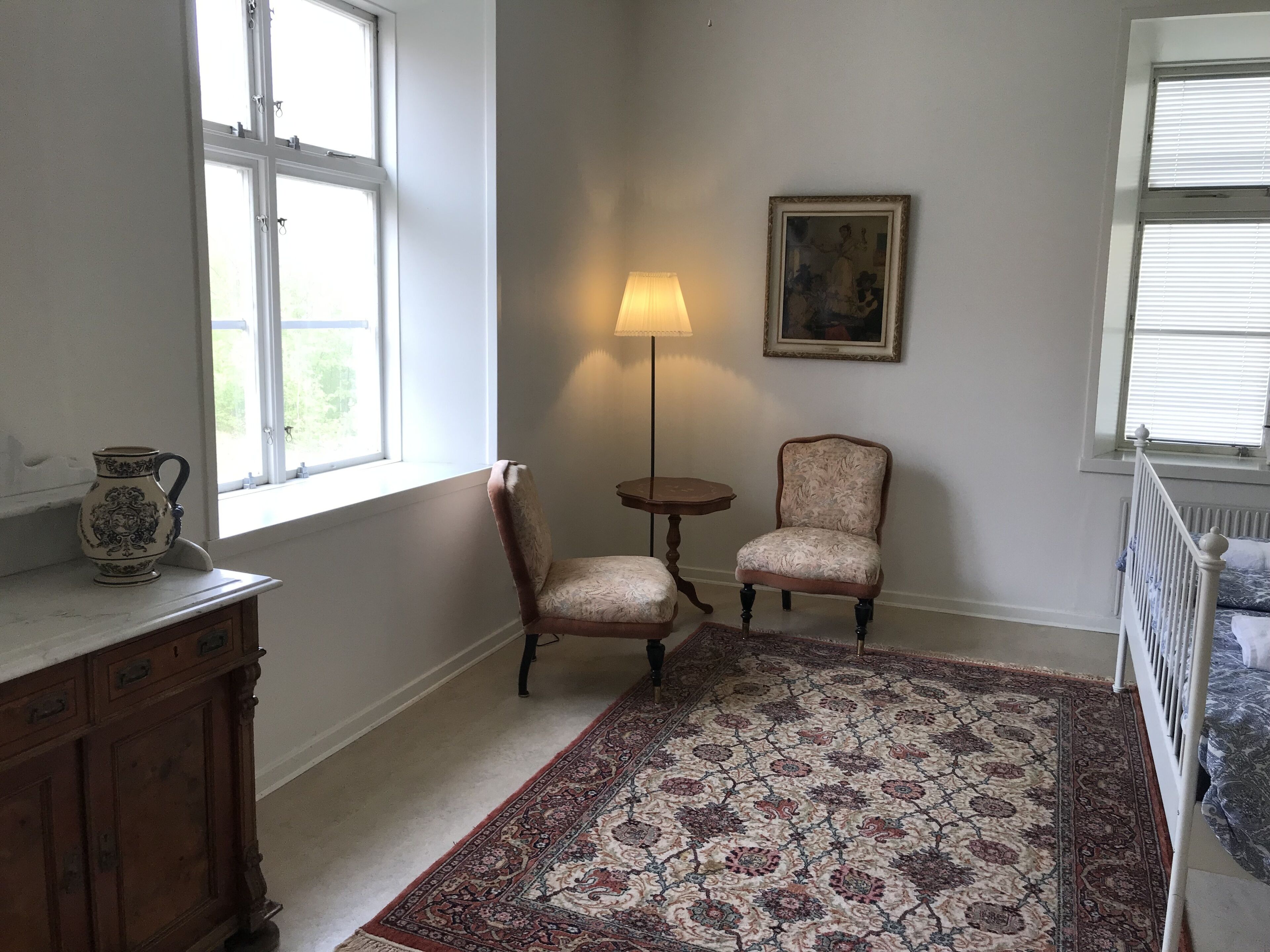 Ett rum med en mönstrad matta, en lampa, två stolar, ett skåp och en tavla på väggen.