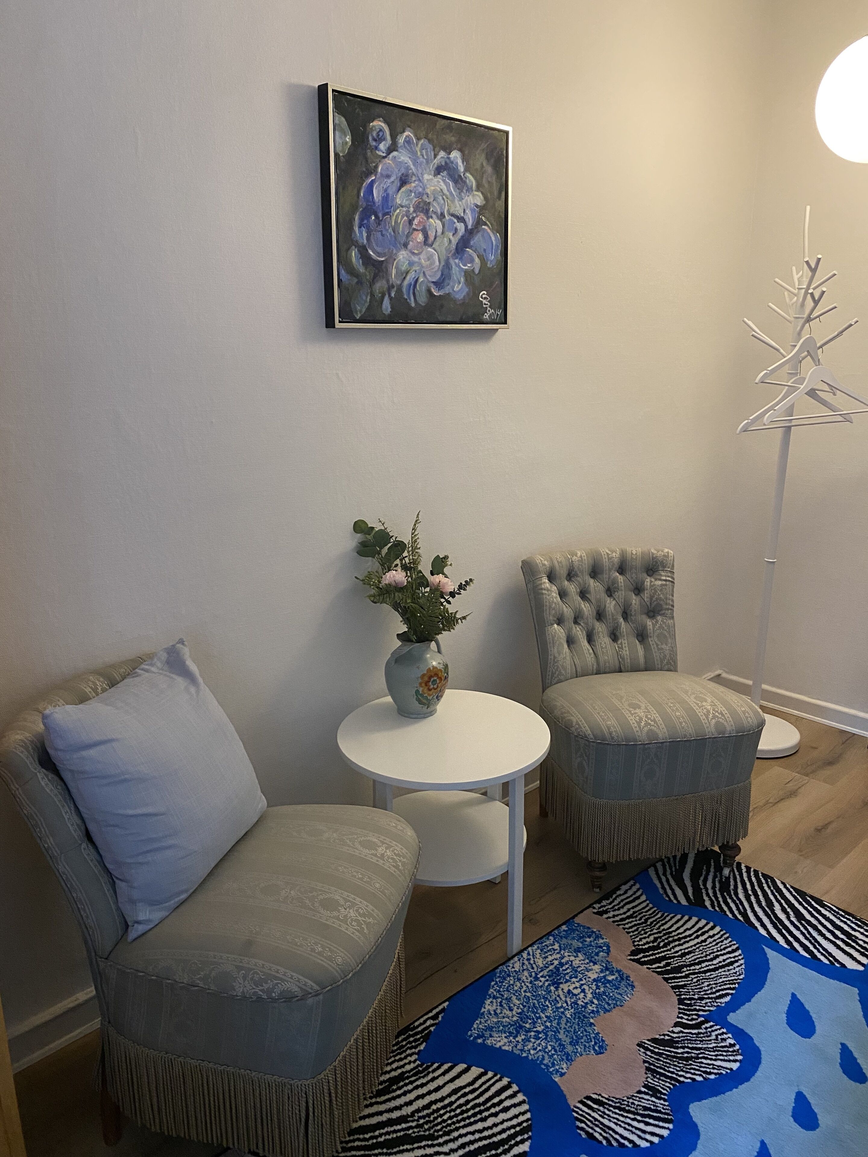 A room with two grey upholstered chairs, a white round table with a vase of flowers, a blue patterned rug, and a painting on the wall.