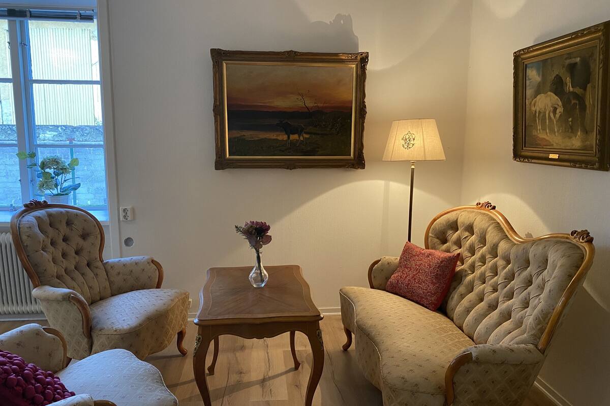 A living room with two armchairs, a small table with a vase, a floor lamp, and two paintings on the wall.