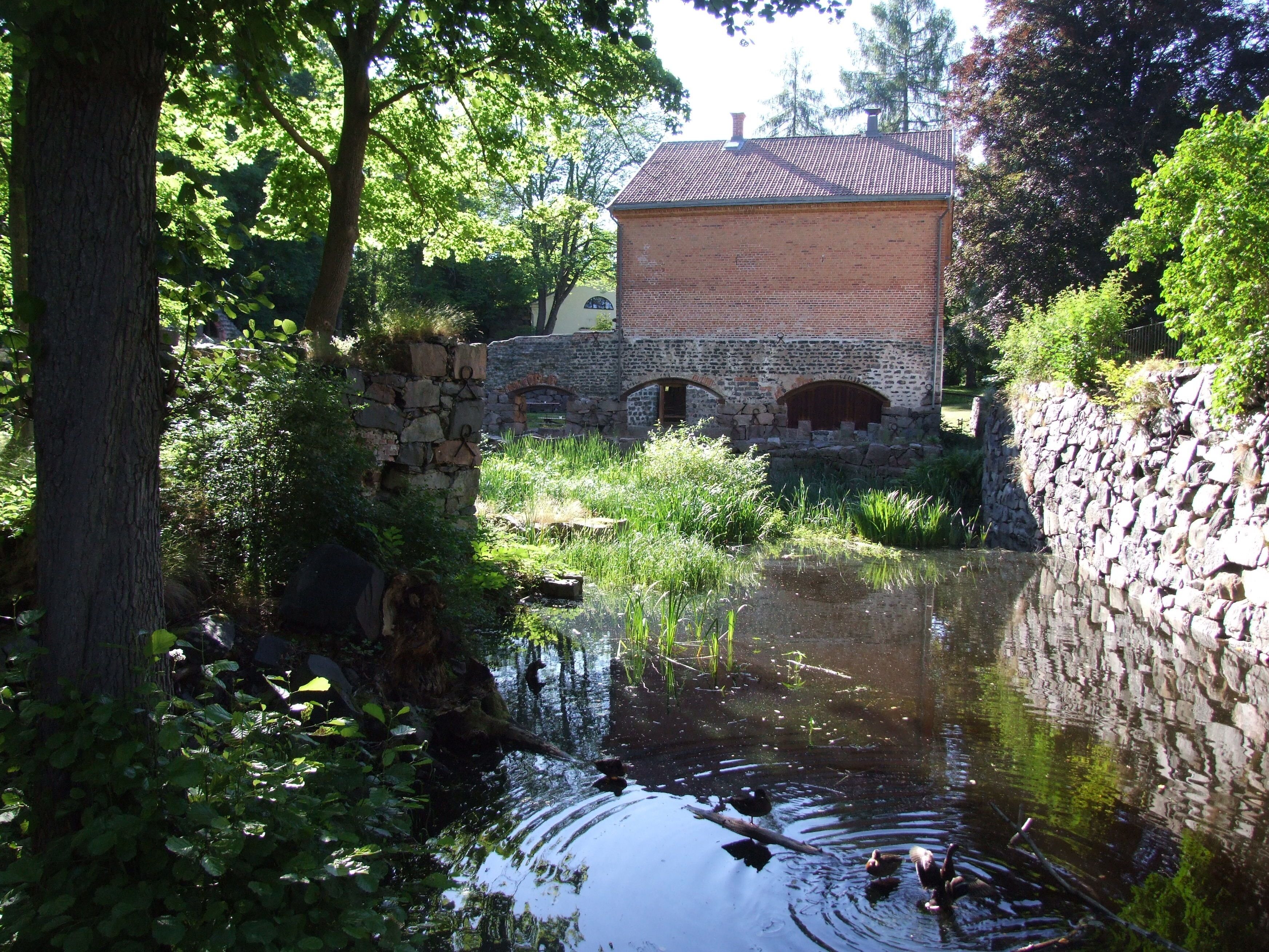 Un bâtiment rustique en pierre, doté d’un toit en tuiles, entouré d’une végétation luxuriante et bordé d’une étendue d’eau paisible.