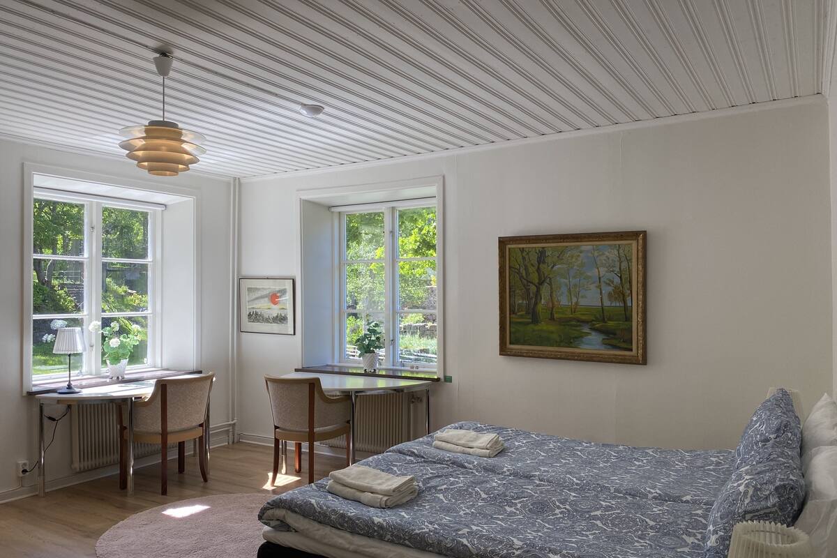 A bedroom with a bed, a desk, and a chair. There are windows and a painting on the wall.