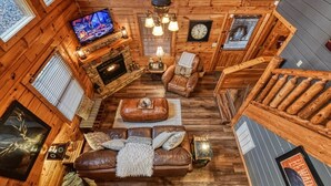 Interior de una cabaña de troncos con chimenea, sofá de cuero, paredes de madera, un televisor y un reloj.