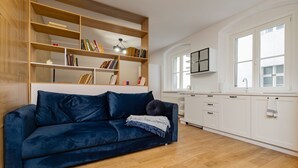 Una sala de estar moderna con un sofá azul oscuro, una estantería de madera y una cocina blanca con una ventana.