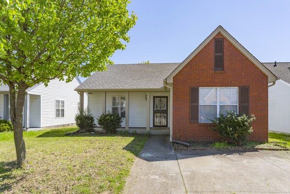 A single-story brick house with a front porch, a garage, and a small front yard with a tree and shrubs.