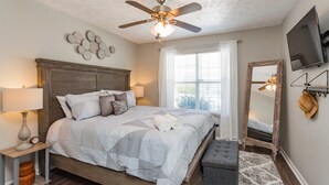 A bedroom with a wooden bed, a ceiling fan, a large window with curtains, a bedside table with a lamp, and a mirror on the wall. - Charming 3bdr home 10 min to downtown! (Nashville)