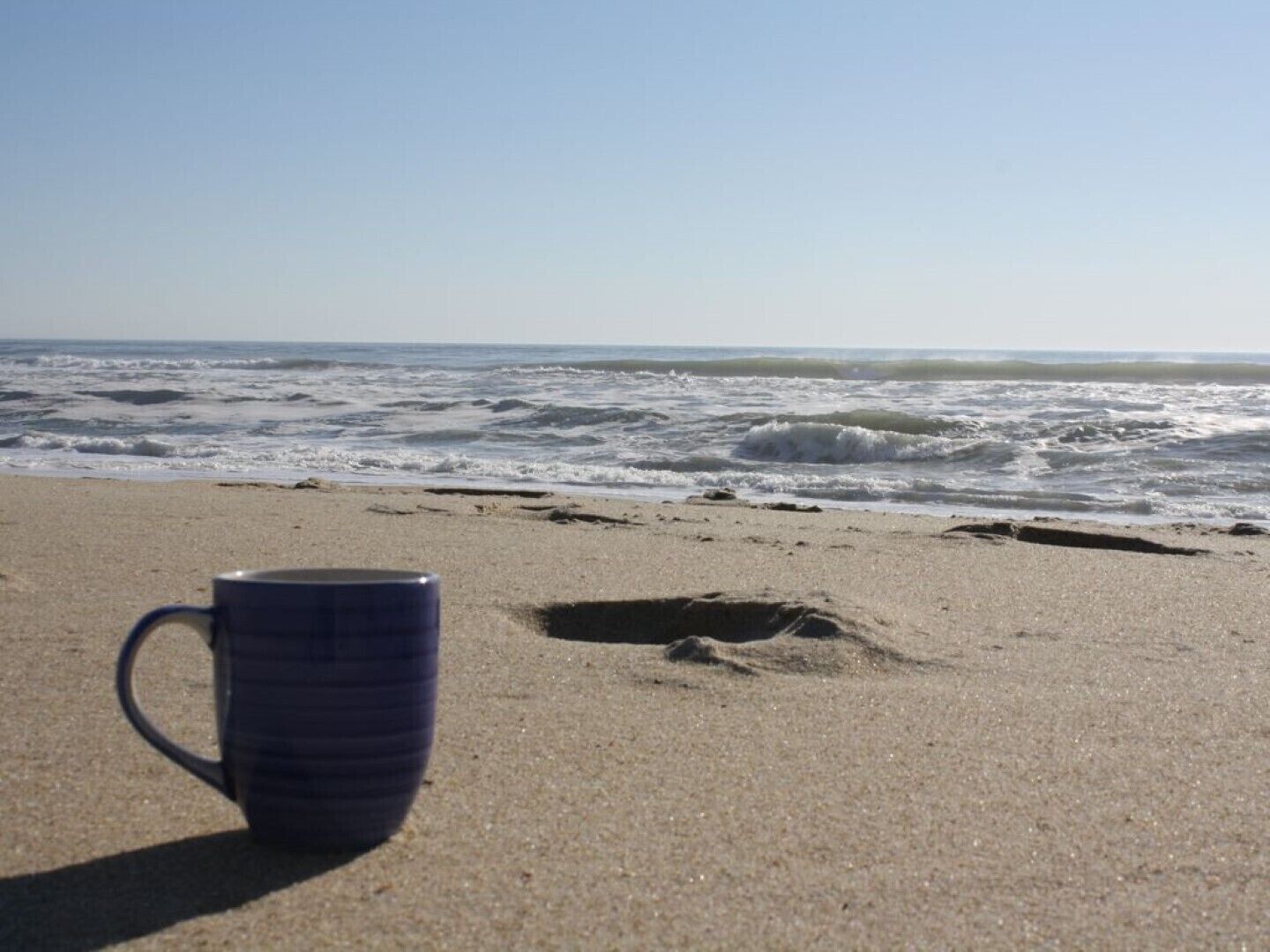 A beach scene with a single blue mug on the sand, waves in the background, and a clear sky.