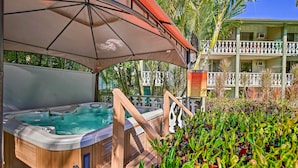 A spa pool area with a wooden hot tub under a large umbrella, surrounded by lush greenery and a building with balconies in the background. - Kailua-Kona Studio - Walk to Hale Halawai Park! (Kailua-Kona)