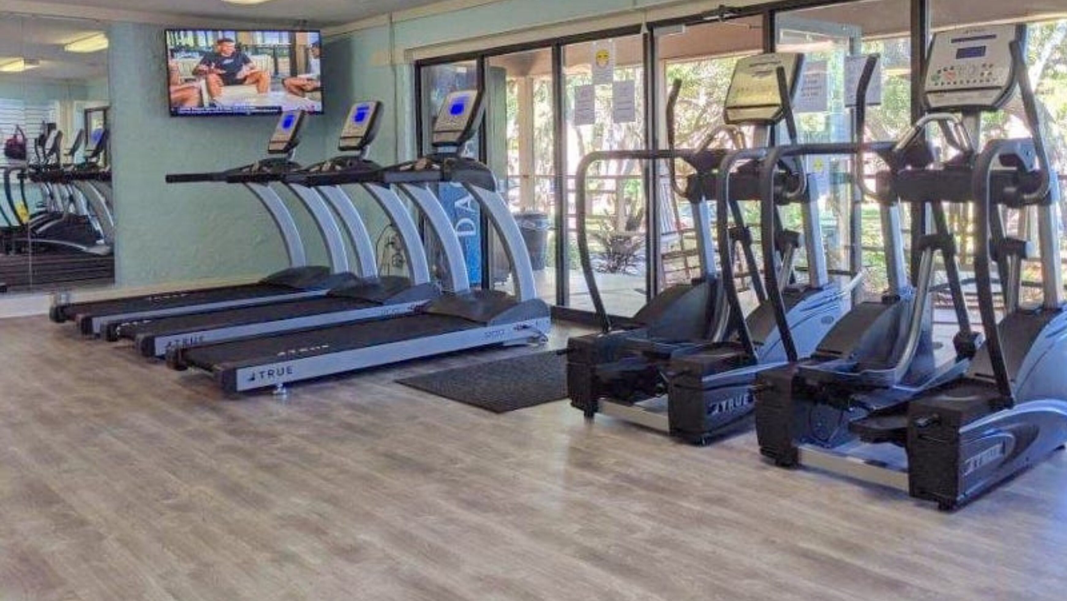 Un gimnasio con cintas de correr, elípticas y una pantalla de televisión mostrando un programa de ejercicios.