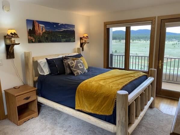 A bedroom with a wooden bed, a nightstand, a painting of a mountain landscape, and a view of a green field through a glass door.