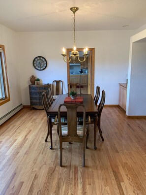 Een eetkamer met een houten tafel, zes stoelen, een kroonluchter, een klok en uitzicht op de keuken.
