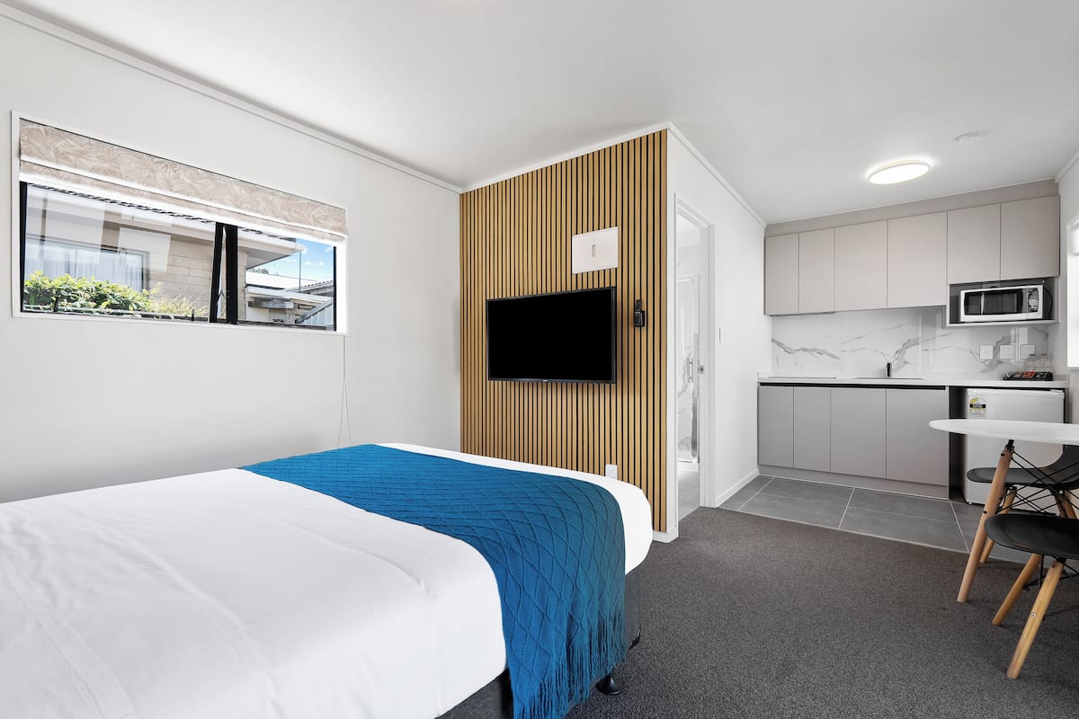 A modern hotel room with a bed, a TV, a small kitchenette, and a window with blinds.