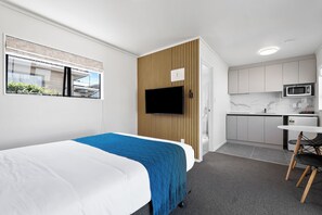 A modern hotel room with a bed, a TV, a small kitchenette, and a window with blinds.