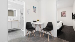 A modern, minimalist room with a white wall, a round table, two black chairs, a small white table with a vase of flowers, and a bed with a dark headboard. - Summer Inn Motel (Orewa)