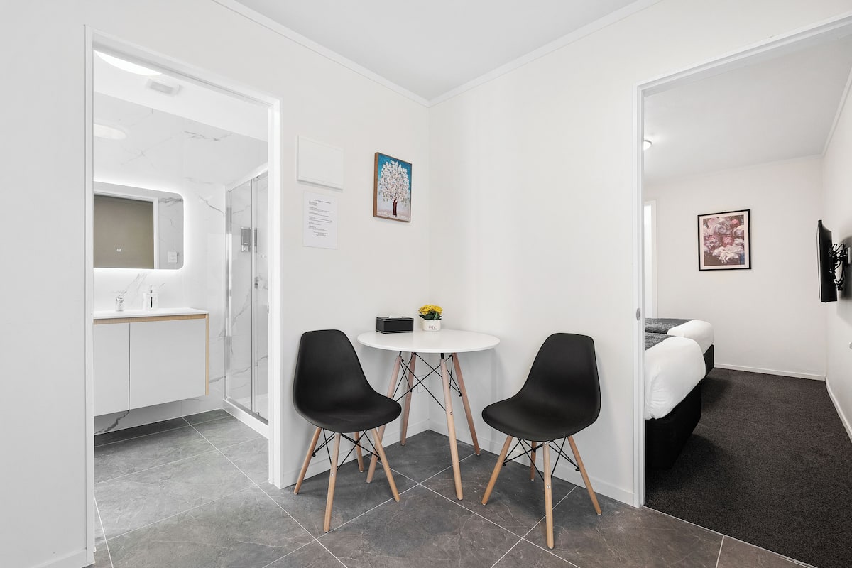 A modern, minimalist room with a white wall, a round table, two black chairs, a small white table with a vase of flowers, and a bed with a dark headboard.