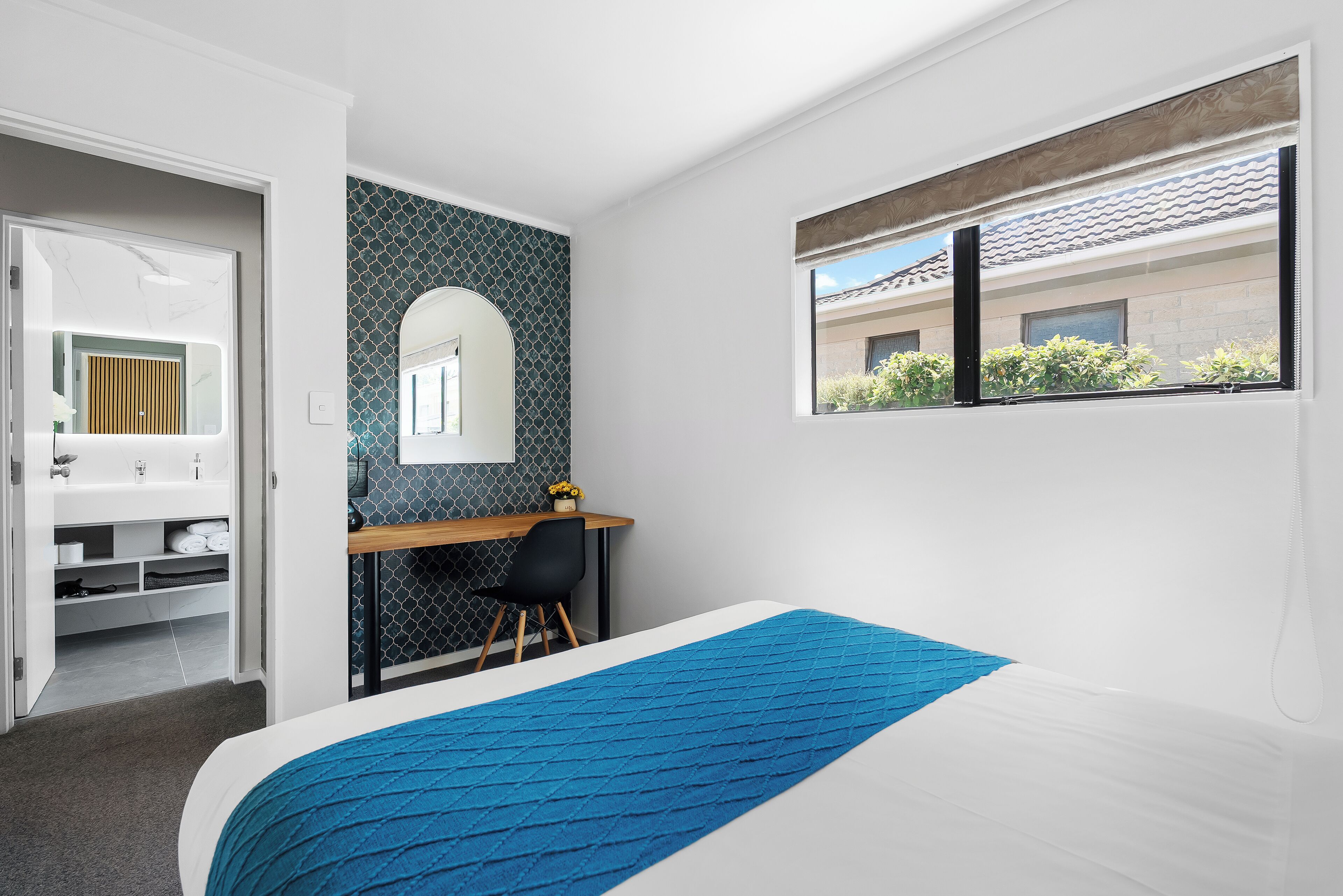 A bedroom with a bed, a desk, a chair, a mirror, and a window with a view of buildings.