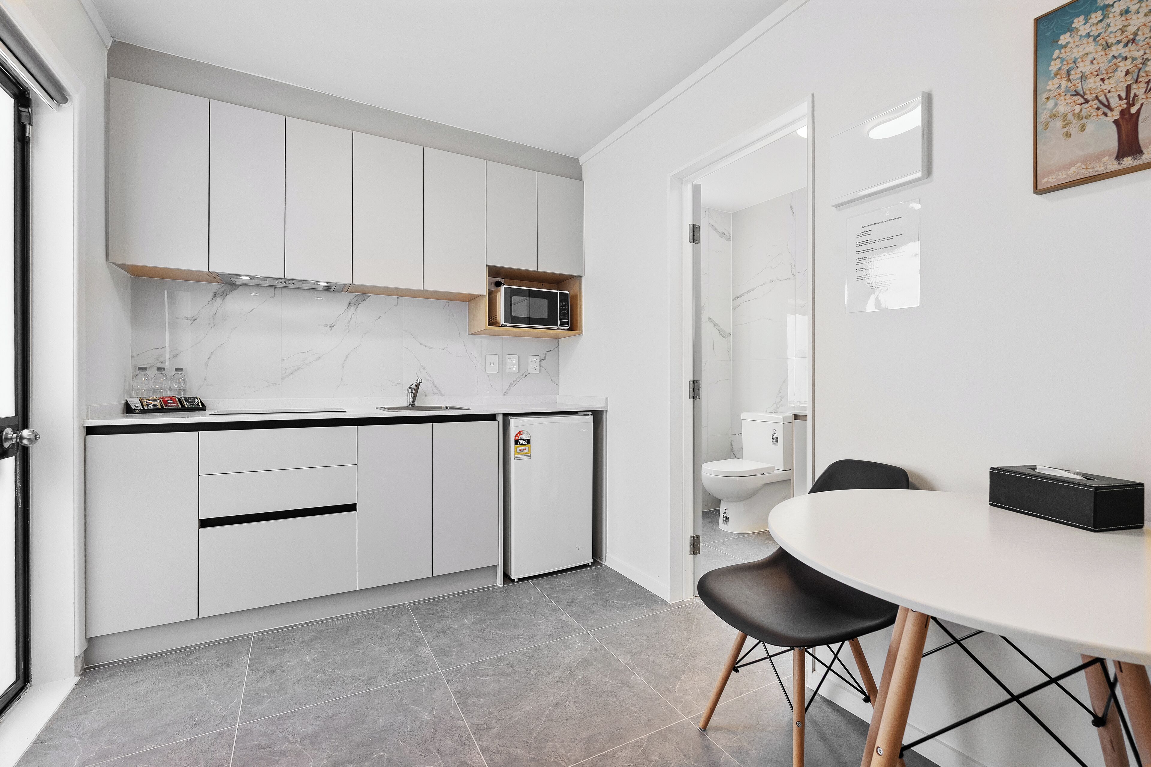 A modern kitchen with white cabinets, a microwave, and a refrigerator. There is a dining table with chairs and a painting on the wall.