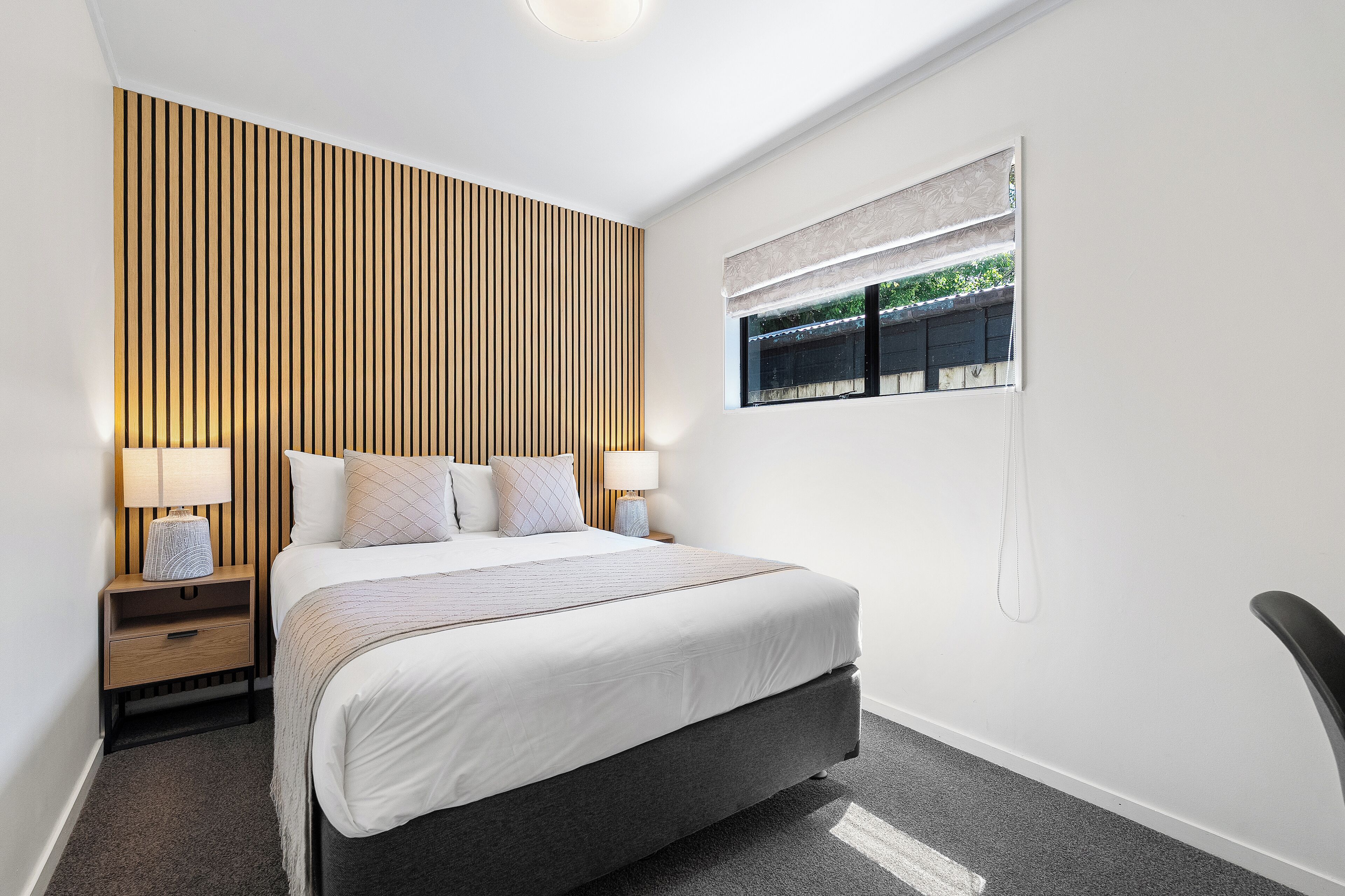 Una habitación de hotel moderna con una cama grande, panel de pared de madera, lámparas de noche y una ventana con persianas.