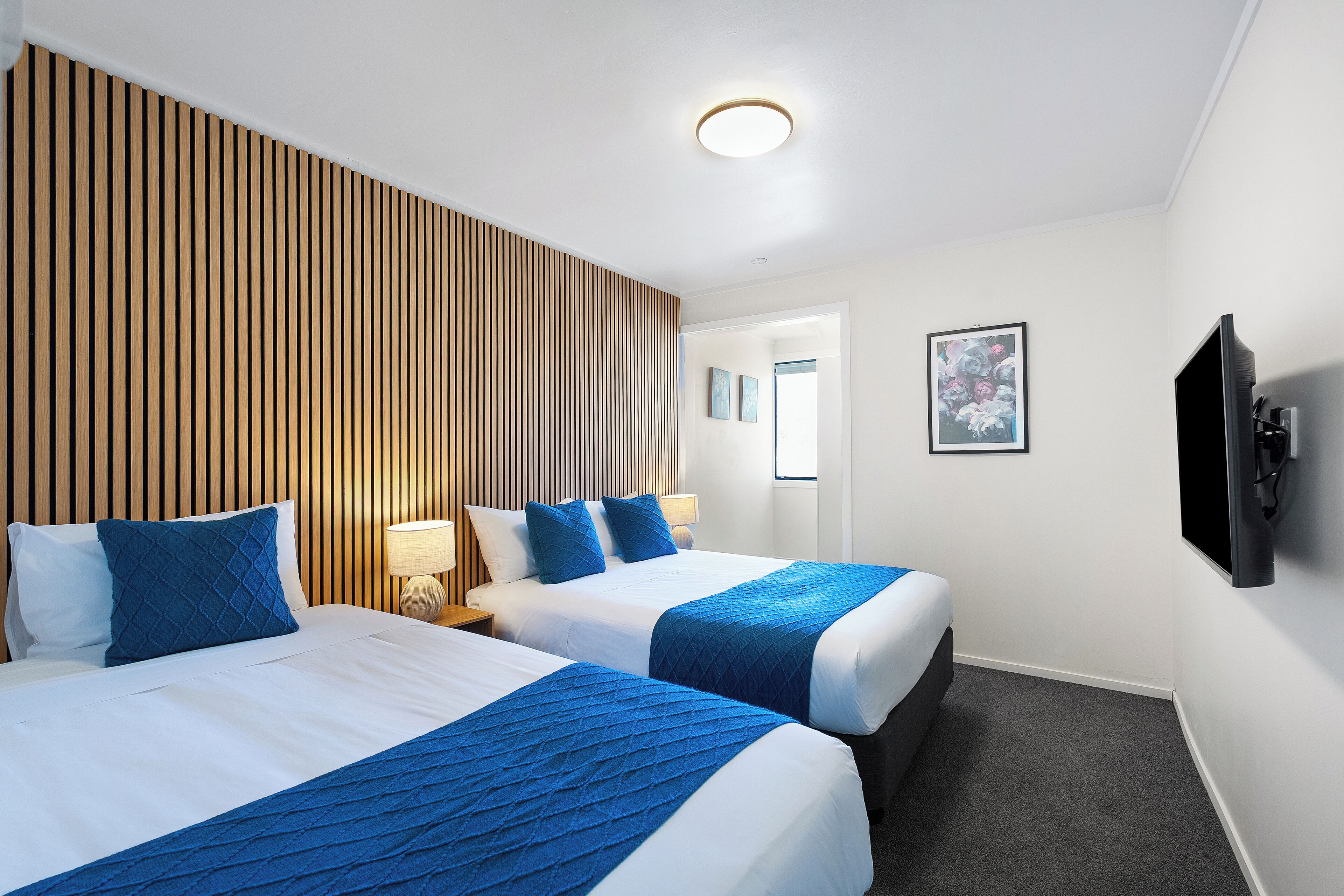 A hotel room with two beds, a wooden slat headboard, a flat-screen TV, and a framed picture on the wall.