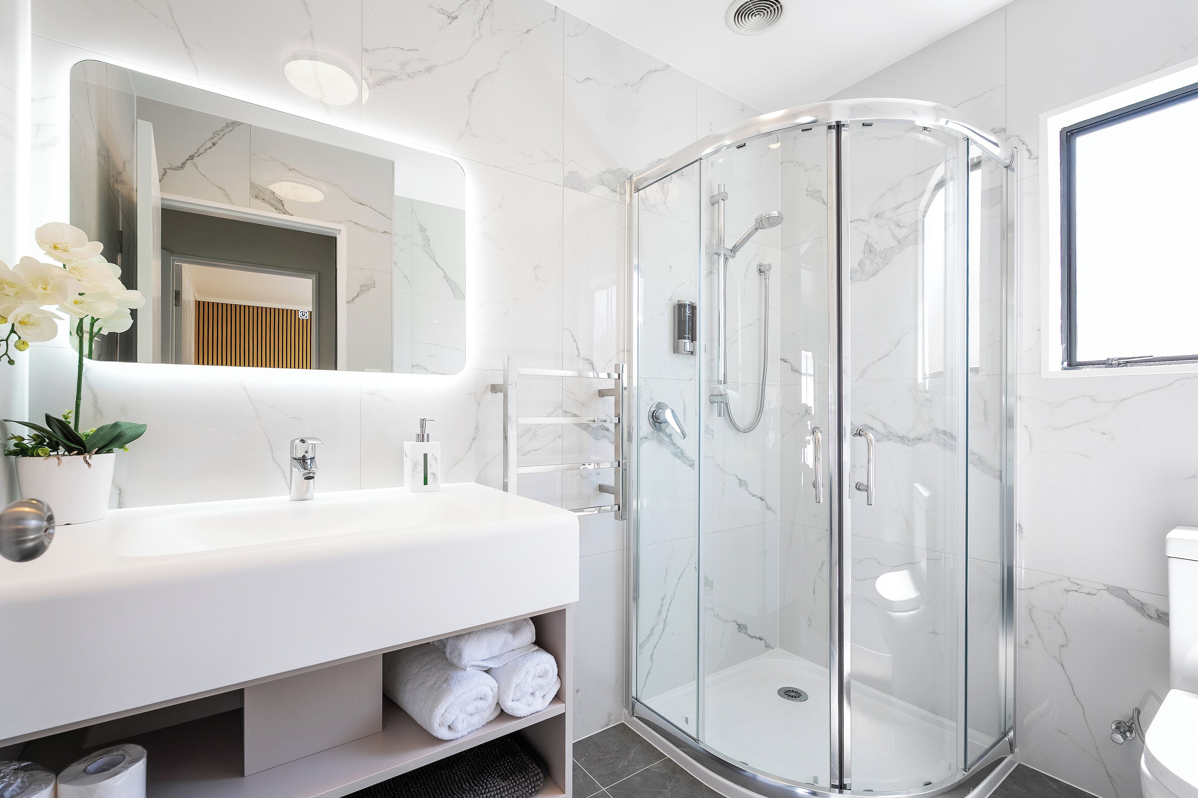 Un baño moderno con lavamanos de mármol blanco, ducha con cerramiento de vidrio y un espejo grande.