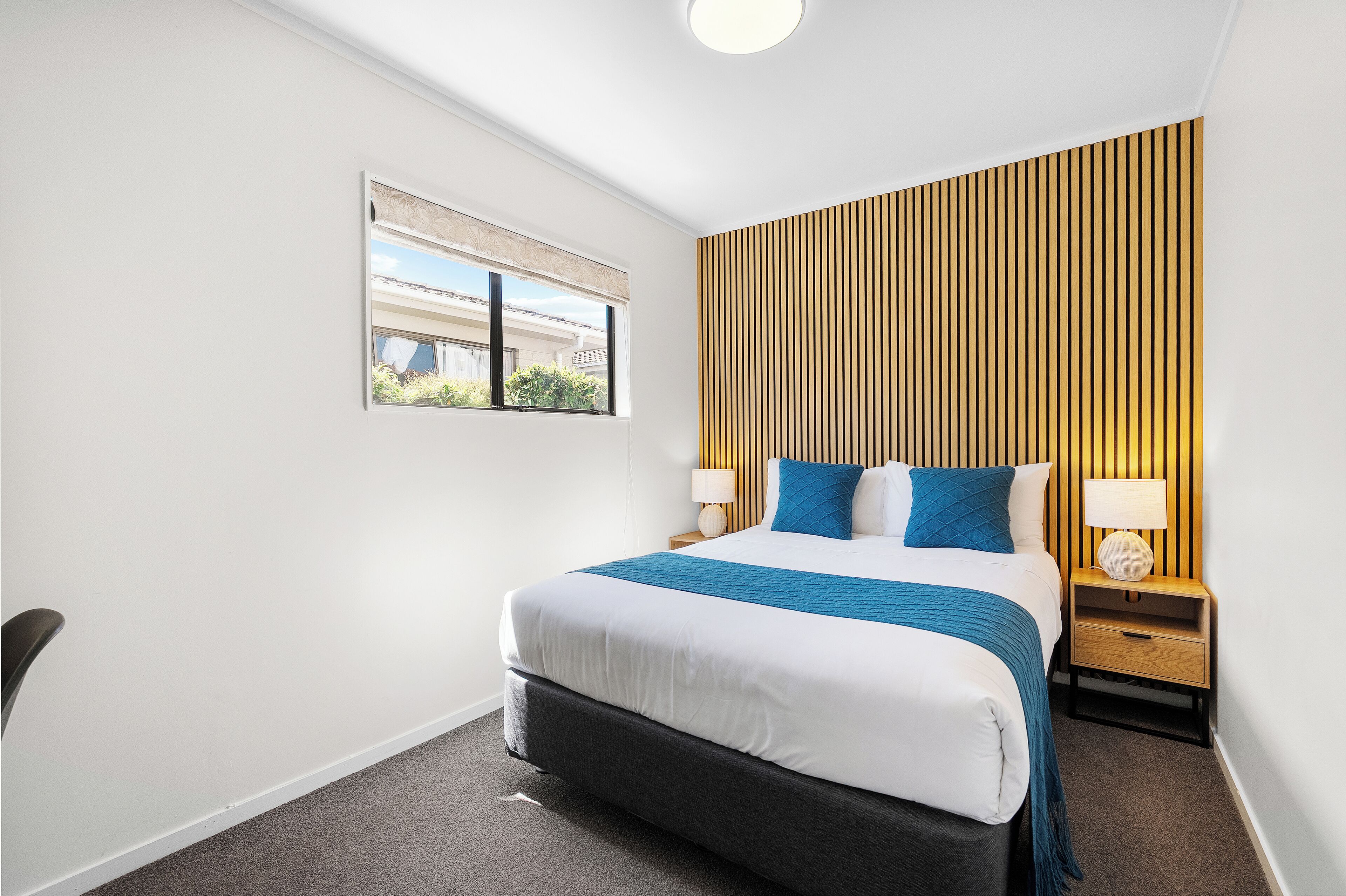 A hotel room with a large bed, a nightstand with a lamp, a window with a view of buildings, and a wooden panel wall.