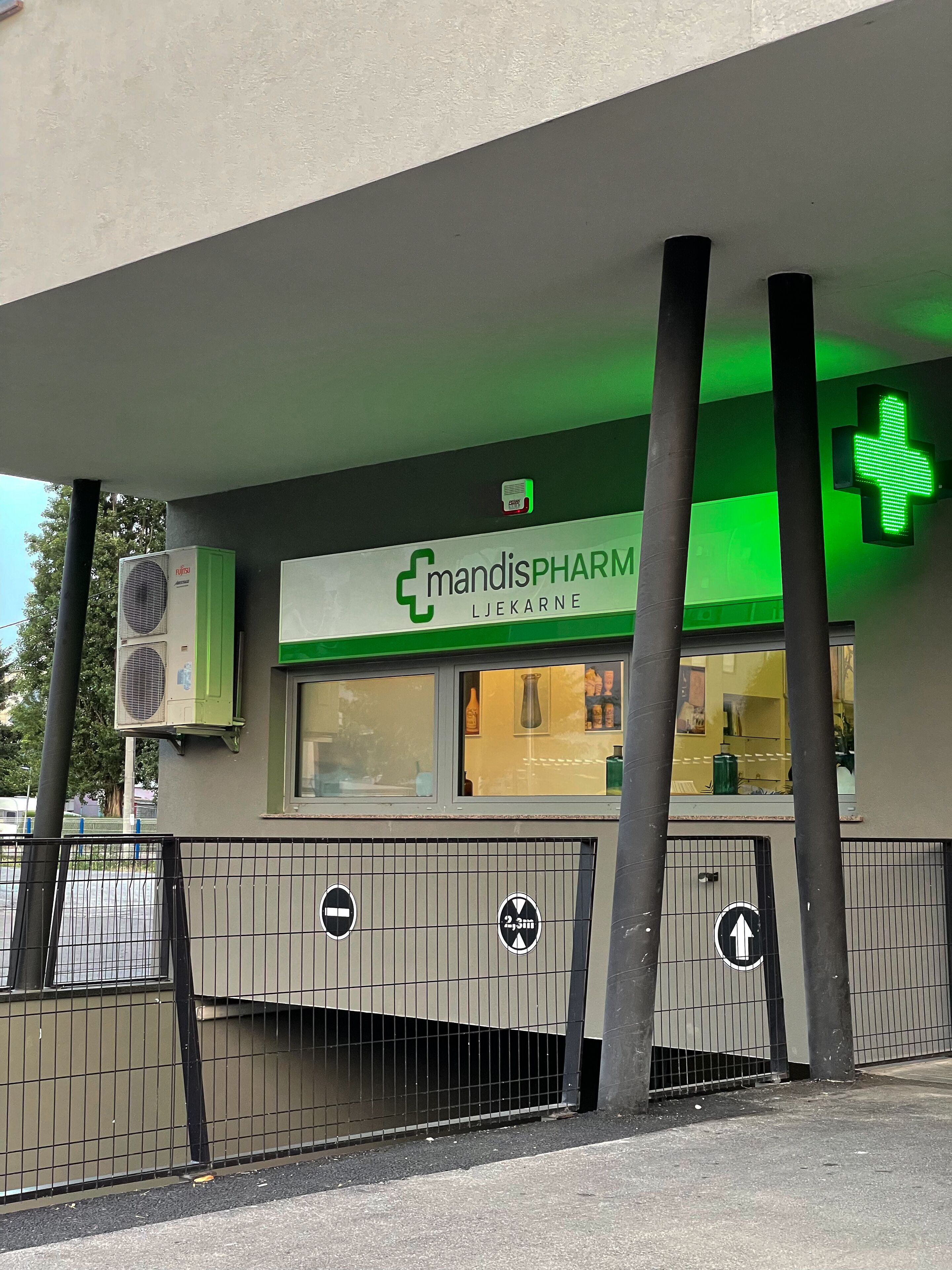 Una farmacia con un letrero de cruz verde, unidades de aire acondicionado y una entrada protegida por una reja.