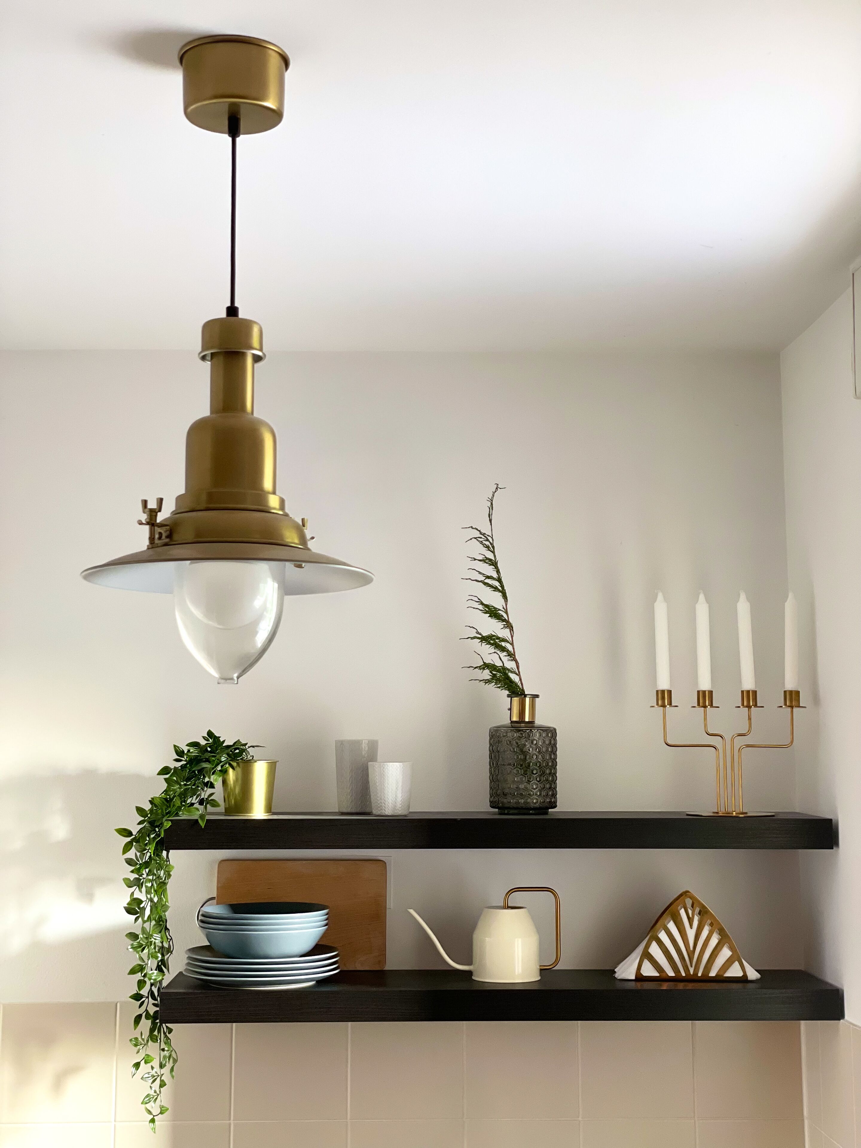 A kitchen with a hanging brass light fixture, a potted plant, a candle holder, and a stack of plates on a black shelf.