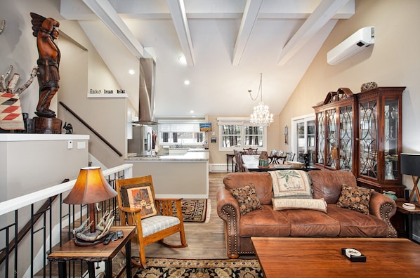 A spacious living room with a brown leather sofa, wooden side table, and a chandelier. A dining area with a table and chairs is visible in the background.