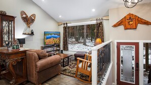 Un salón acogedor con chimenea, televisor y vistas a un bosque nevado a través de la ventana.