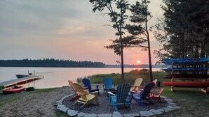 Un entorno junto al lago con una zona de estar circular rodeada de sillas Adirondack, un muelle con un bote y una pila de canoas.