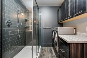 A modern bathroom with a glass-enclosed shower, a built-in washing machine, and dark tiled walls.