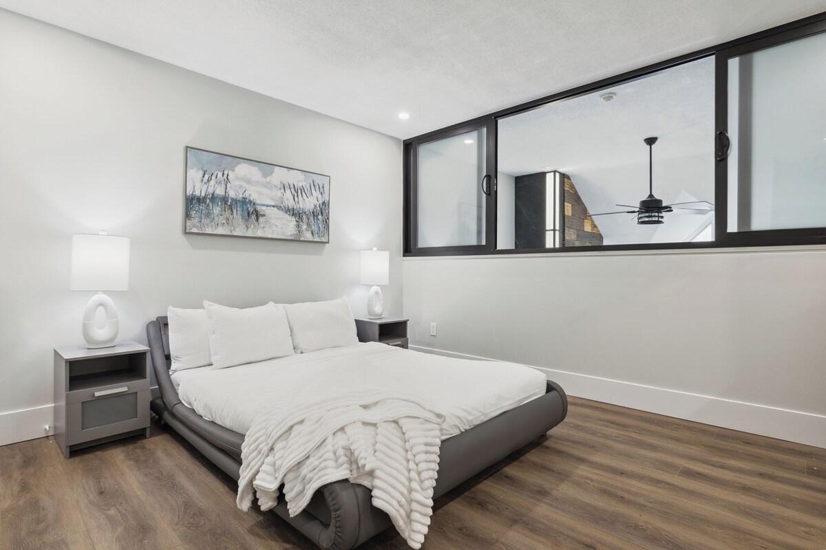 A modern bedroom with a large bed, bedside tables, a painting, and a view through a window.