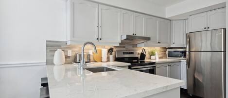 A modern kitchen with white cabinetry, a stainless steel refrigerator, a built-in oven, and a marble countertop.