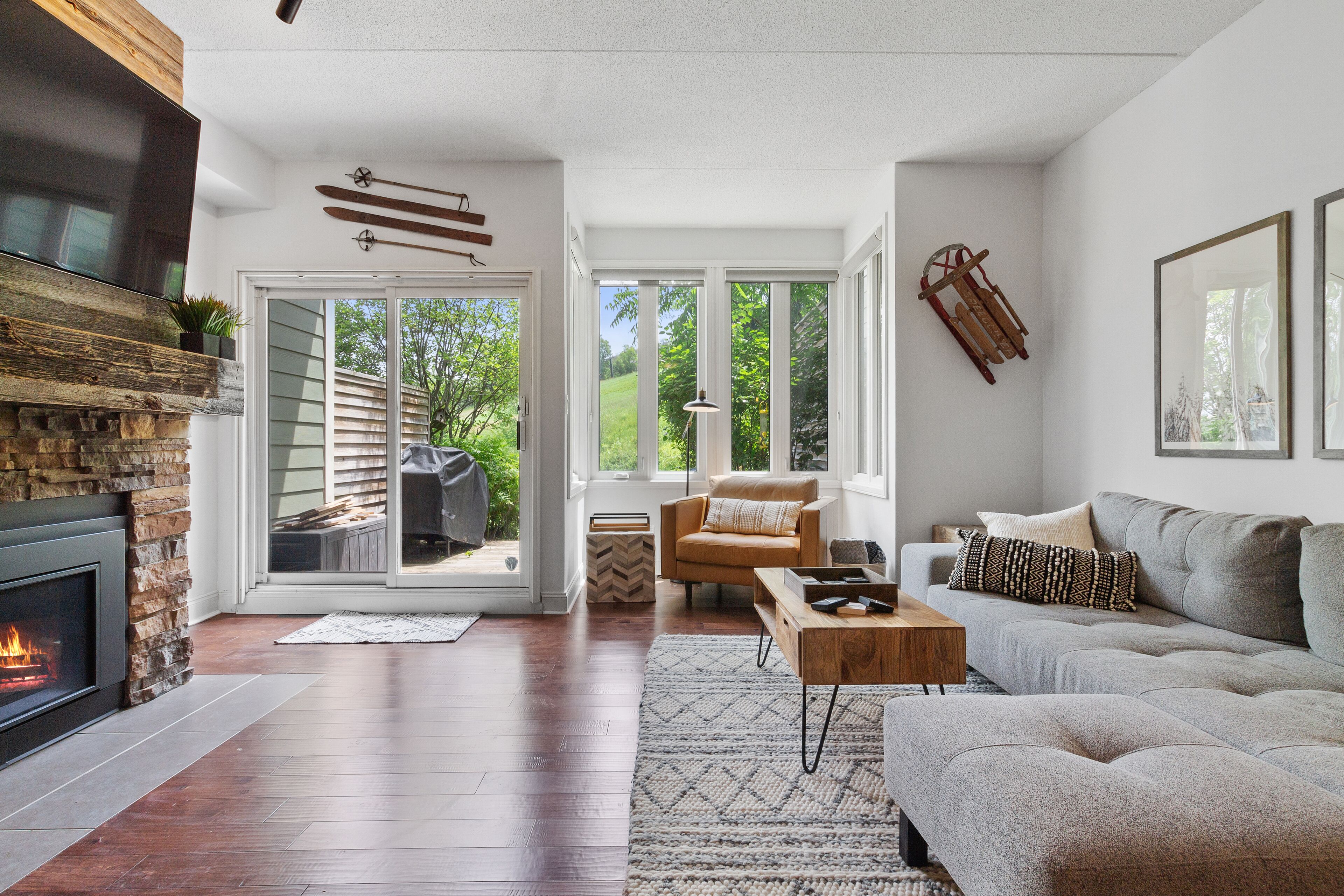 A modern living room with a fireplace, a grey sofa, a wooden coffee table, and a large window offering a view of the outdoors.