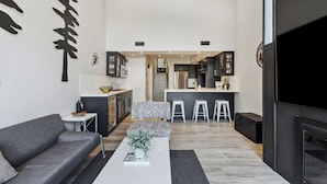 Un salon moderne avec un canapé gris, une table basse blanche et un pouf à motifs. On aperçoit, au fond, une cuisine avec des meubles sombres et un îlot central.