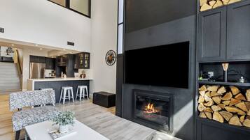 A modern living room with a fireplace, a kitchen in the background, and a dining area with a white table and patterned chairs.