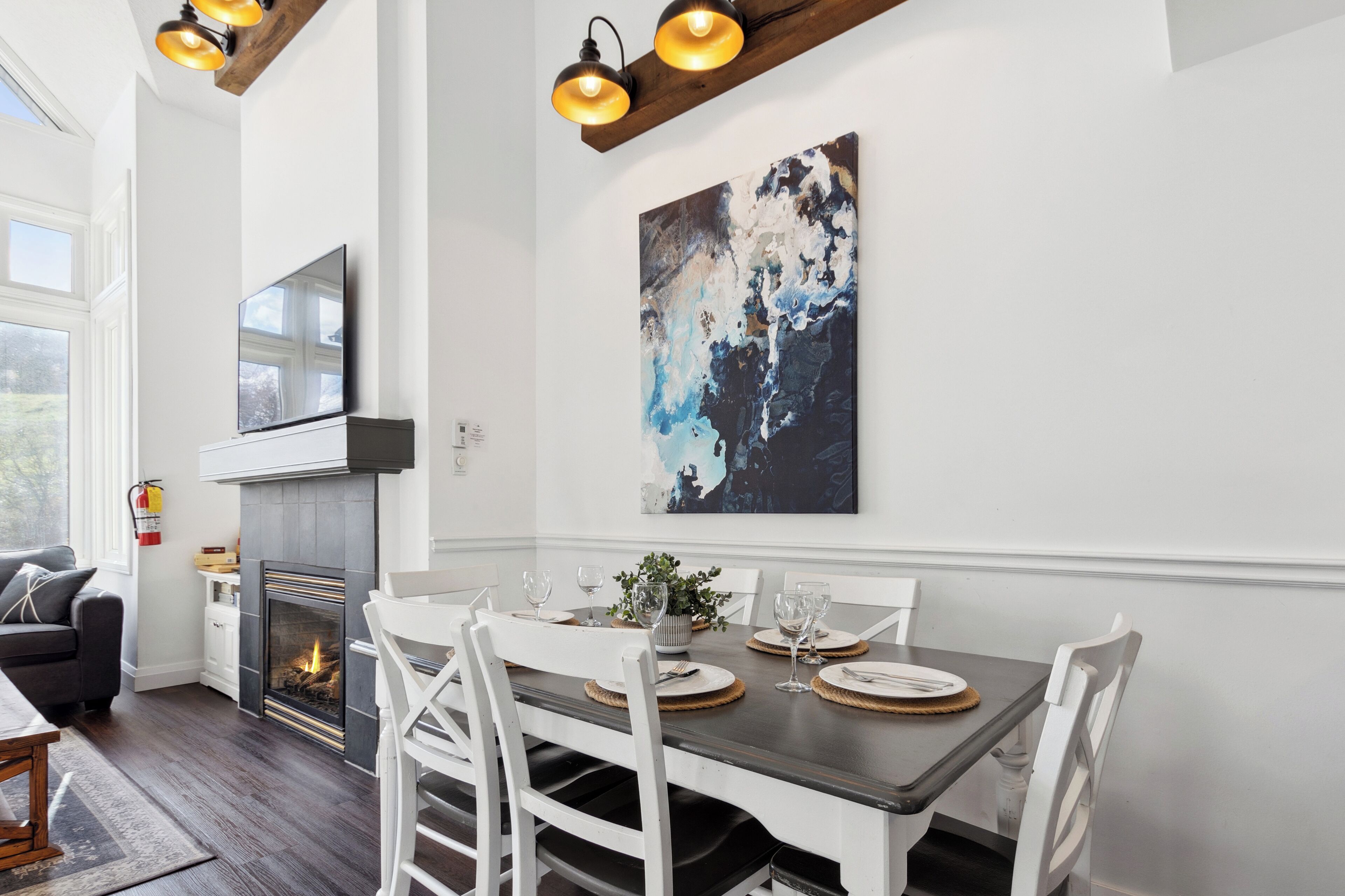 A dining area with a fireplace, white chairs, a table set for four, and a large abstract painting on the wall.