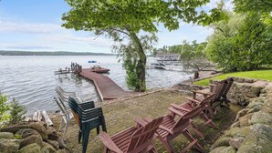 A lakeside deck with Adirondack chairs, a dock extending into the water, and a building with a dock in the background. - Twilight Cove: "Beautiful Sunset Views on Canandaigua Lake!" (Canandaigua)
