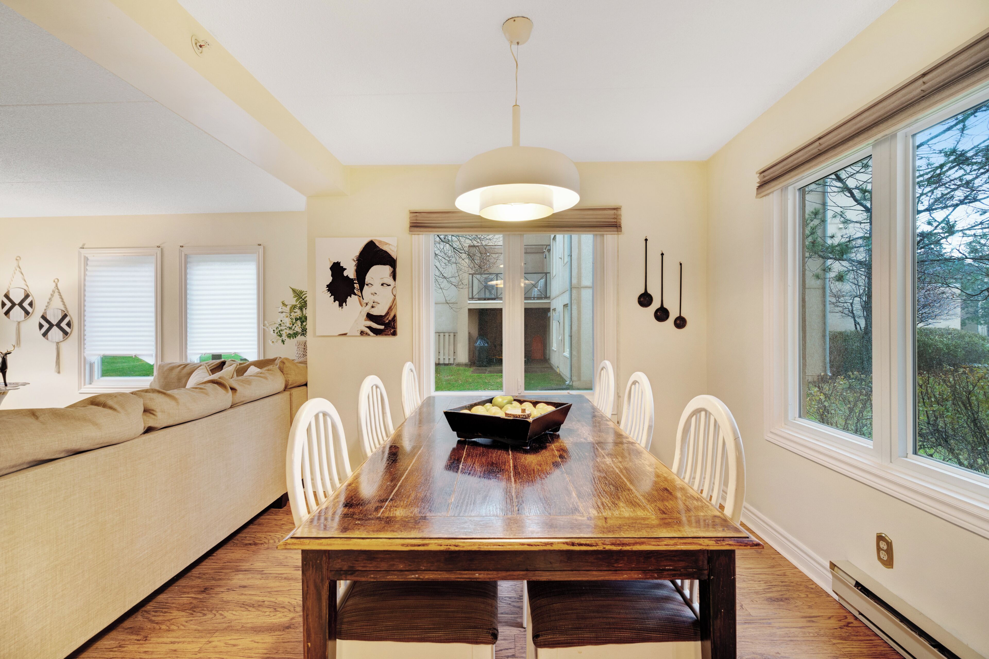 A dining room with a wooden table, white chairs, and a bowl of fruit. There is a sofa with cushions and a painting on the wall.