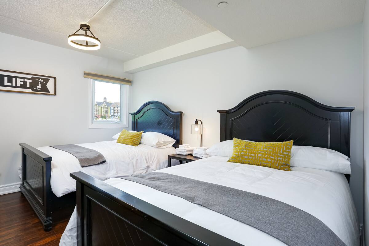 A hotel room with two beds, a window with a city view, a lift sign, and a modern ceiling light.
