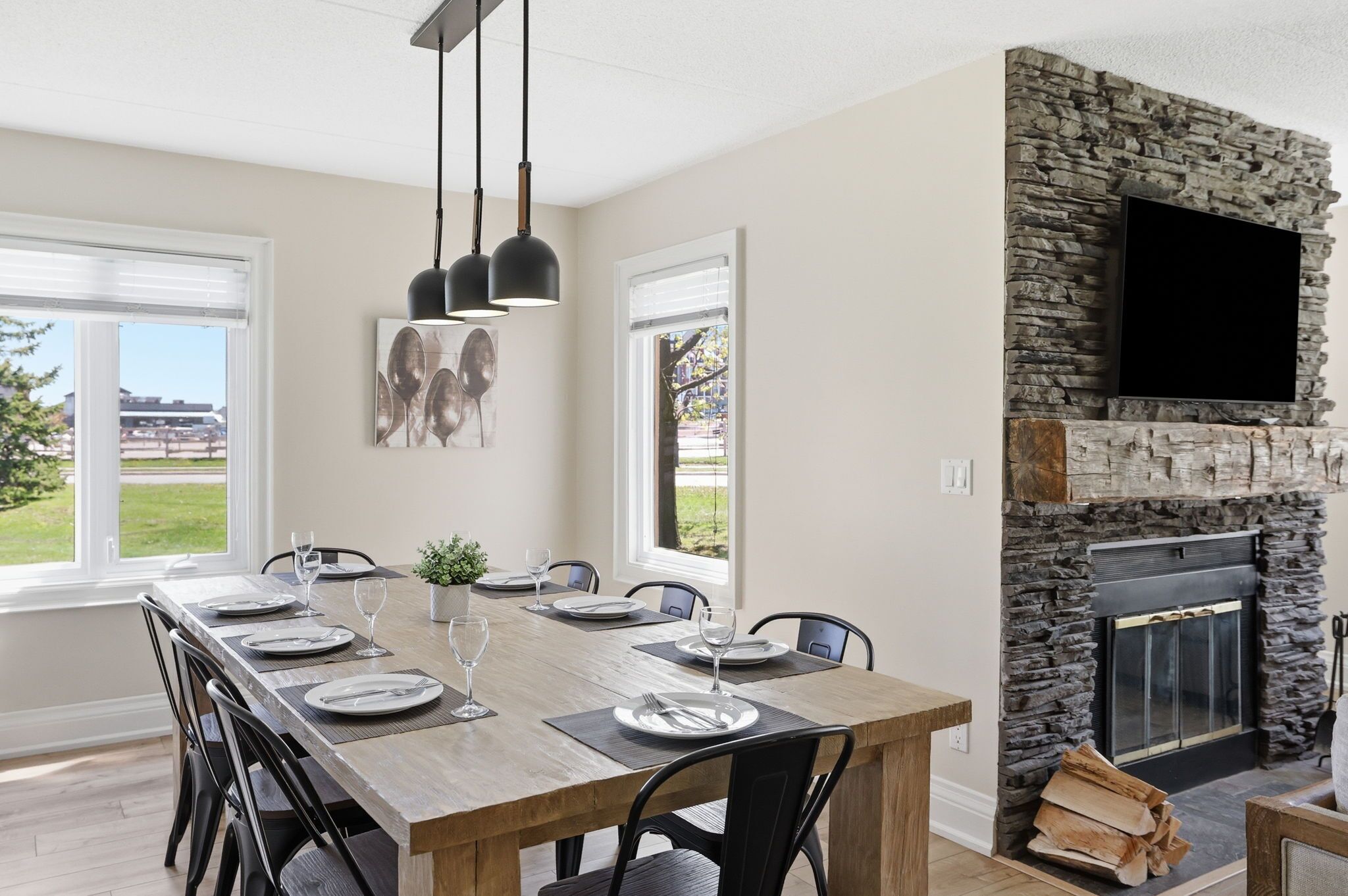 A dining area with a long wooden table set for six, black chairs, and pendant lights. A stone fireplace with a flat-screen TV is visible.