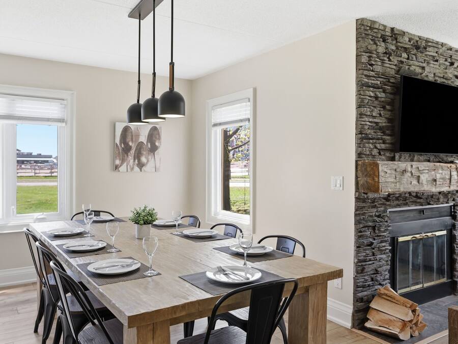 A dining area with a long wooden table set for six, black chairs, and pendant lights. A stone fireplace with a flat-screen TV is visible.
